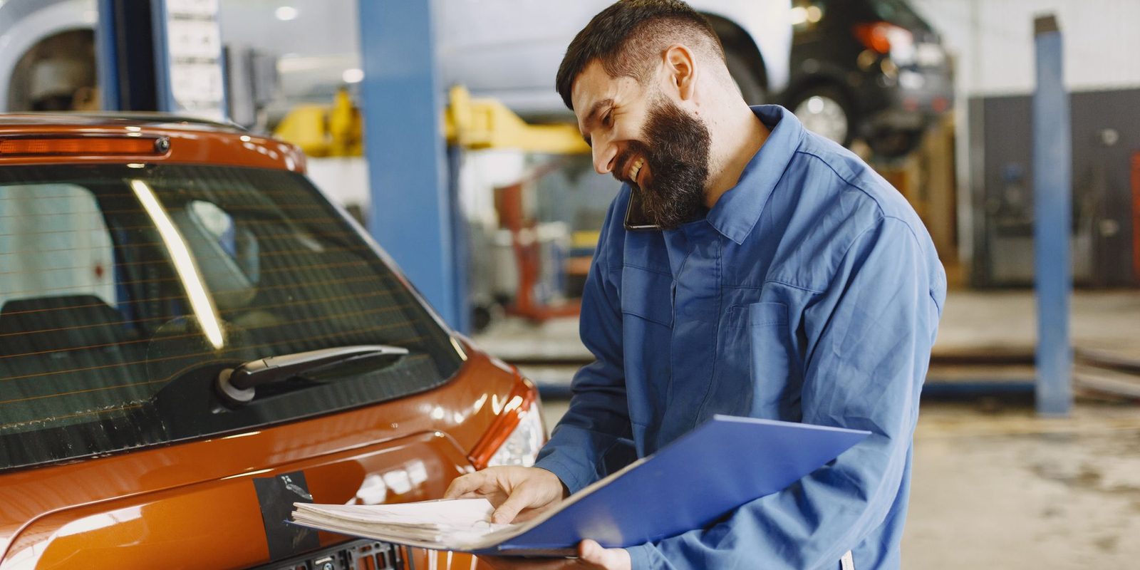 técnico de macacão azul faz anotações numa prancheta apoiada no carro, dentro de uma oficina