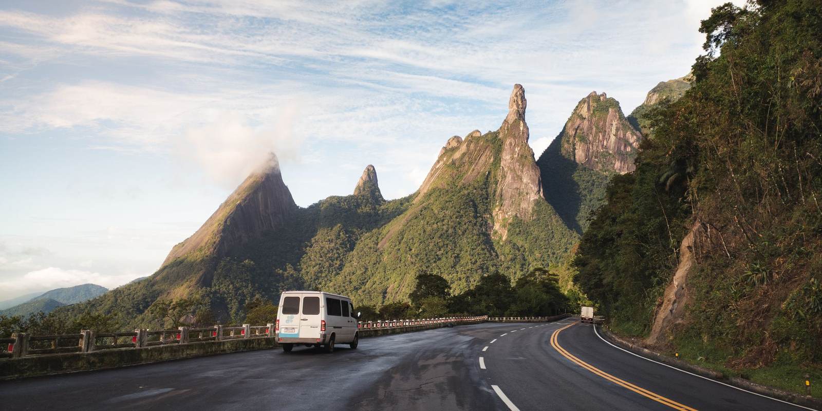 van branca em curva de estrada de serra, com o pico Dedo de Deus e outros paredões de granito ao fundo, na região serrana do Rio de Janeiro